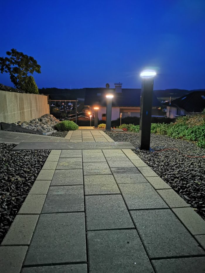 En sti af firkantede fliser fører ned, omkranset af grus og lavt grønt. Moderne, høje Stålstolpe 80x80mm lamper oplyser stien med zquare lys mod aftenhimlen. Et hus dukker op for enden med et træ i silhuet mod den blå nat.