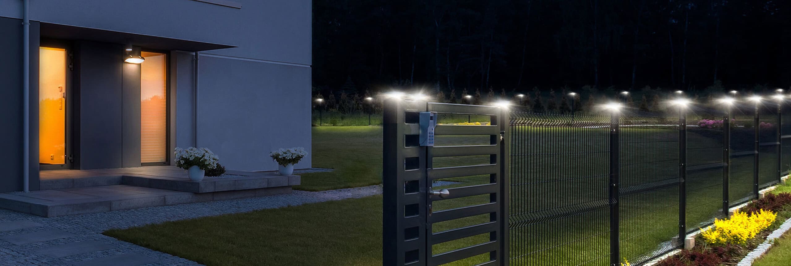 Moderne hus udvendigt med åben port, varmt oplyst indgang og oplyst gangsti. En mørk himmel giver kontrast og fremhæver den pænt trimmede græsplæne og farverige have til højre.