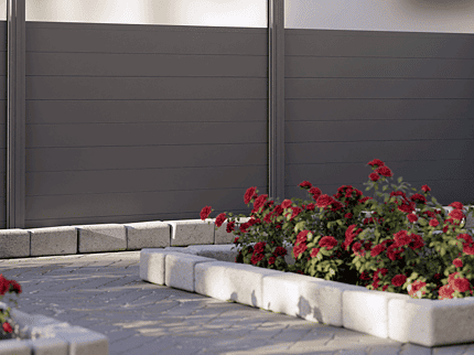 Vidual Snap 140 – Aluminiumshegn med klik-profiler, et moderne gråt privat hegn med matterede glaspaneler øverst, står ved siden af levende røde blomster i et havebed med stenkanter. En asfalteret sti løber langs med, der fører til en bygning og fremhæver naturens blanding med moderne design.