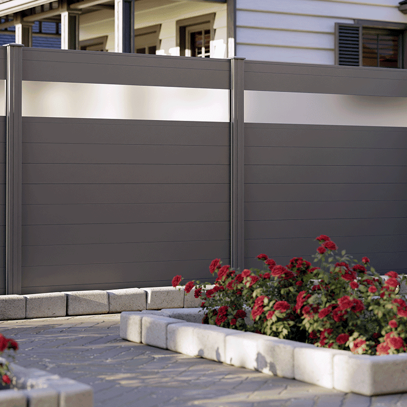 Vidual Snap 140 – Aluminiumshegn med klik-profiler, et moderne gråt privat hegn med matterede glaspaneler øverst, står ved siden af levende røde blomster i et havebed med stenkanter. En asfalteret sti løber langs med, der fører til en bygning og fremhæver naturens blanding med moderne design.