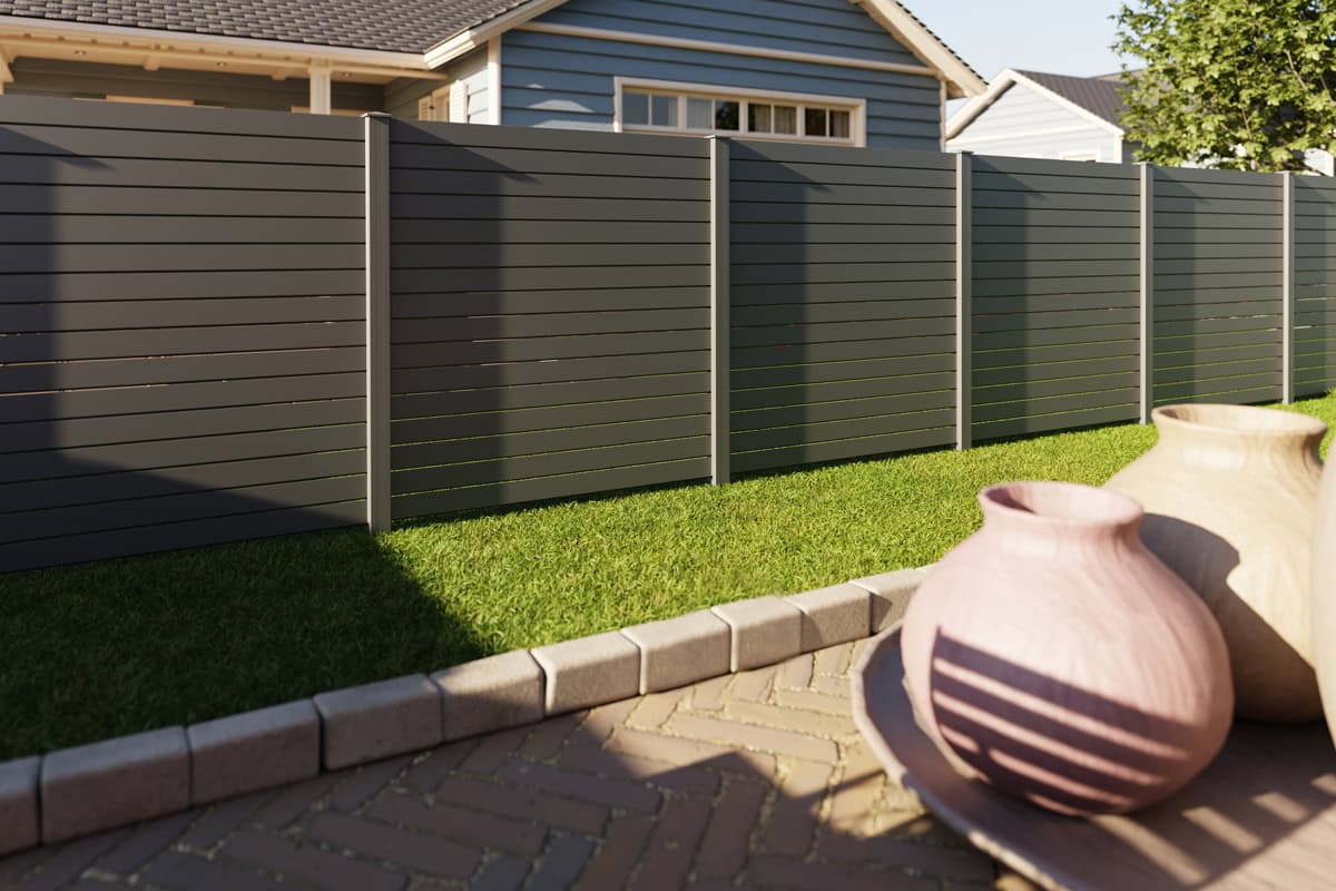 Vidual Pure – Jalousihegn i aluminium med rhombeprofil privatlivshegn står i en baghave med grønt græs, en brolæggerterrasse og vaser. Et lyseblåt hus med hvid trim er synligt bag det moderne grå aluminiumshegn.