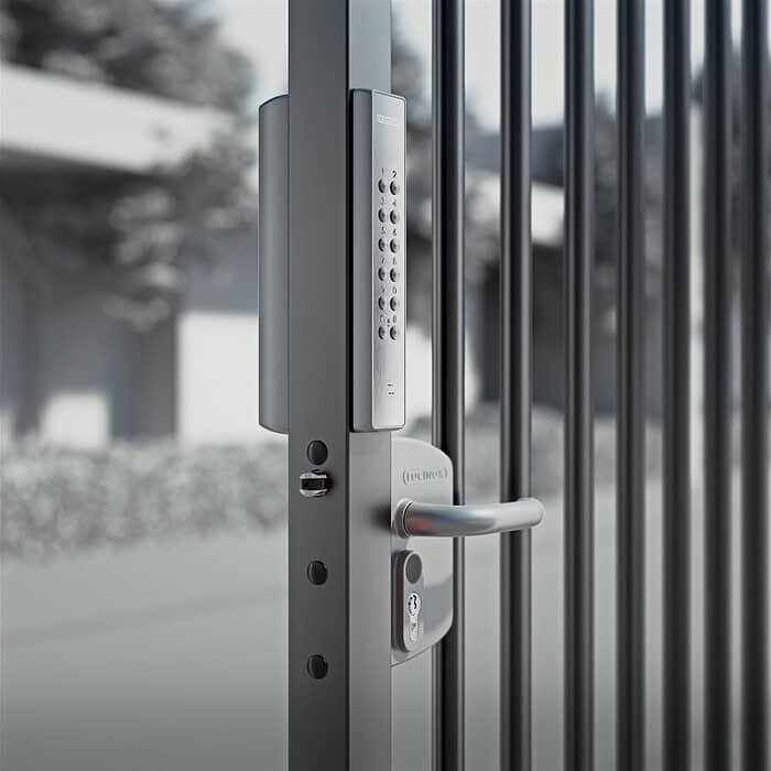 En slank VOLTA - Overflademonteret lås med eksternt batteridrevet tastatur med numerisk tastatur pryder metalporten, sat mod en sløret udendørs scene af træer og bygninger.