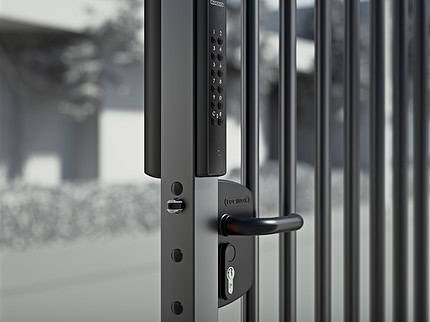 Et nærbillede af den slanke sorte VOLTA elektroniske lås med en overflademonteret lås og et eksternt batteridrevet tastatur, fastgjort til en metalport. Den kunstnerisk slørede baggrund antyder træer og en bygning i udendørs omgivelser.