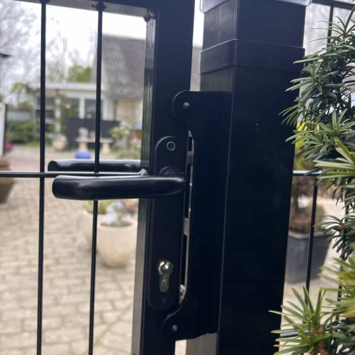 Nærbillede af et sort metalporthåndtag med et Locinox-overflademonteret slutblik/anslag i rustfri stål til indstikslåse på et trådhegn; havegårdsplads og potteplanter blødt sløret i baggrunden.