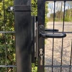 Nærbillede af en sort metalport med håndtag og Locinox-overflademonteret slutblik/anslag i rustfri stål til indstikslåse, nøgler i låsen. Grønne buske, grussti og træer kan ses i baggrunden.