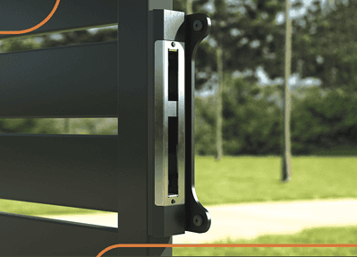 Nærbillede af en Locinox overflademonteret slutblik i rustfrit stål mod en sløret baggrund af grønt græs, træer og en sti. Den slanke, metalliske lås har en lodret rektangulær spalte for vedvarende styrke.