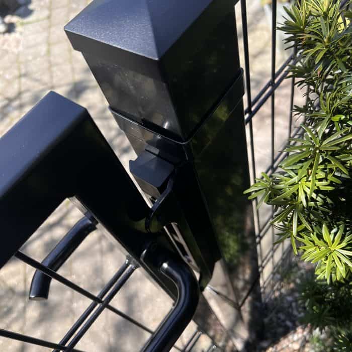 Et nærbillede af et Locinox-overflademonteret slutblik/anslag i rustfri stål til indstikslåse monteret på et trådhegn, omgivet af grønne områder og fortov, med sollys reflekteret fra den rustfri ståloverflade.