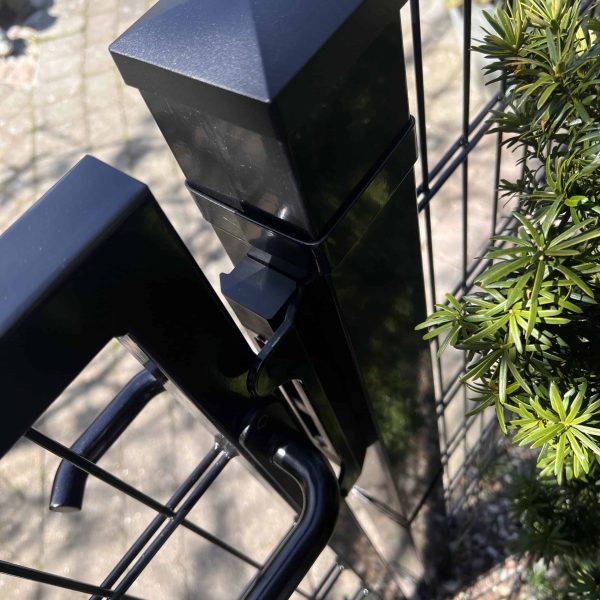 Et nærbillede af et Locinox-overflademonteret slutblik/anslag i rustfri stål til indstikslåse monteret på et trådhegn, omgivet af grønne områder og fortov, med sollys reflekteret fra den rustfri ståloverflade.