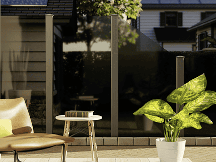 En hyggelig udendørs terrasse har en solbrun loungestol, et lille bord med en bog og en grøn potteplante. Vidual Clear – Glashegn med aluminiumsstolper giver privatliv, mens solpanelklædte huse er synlige i baggrunden.