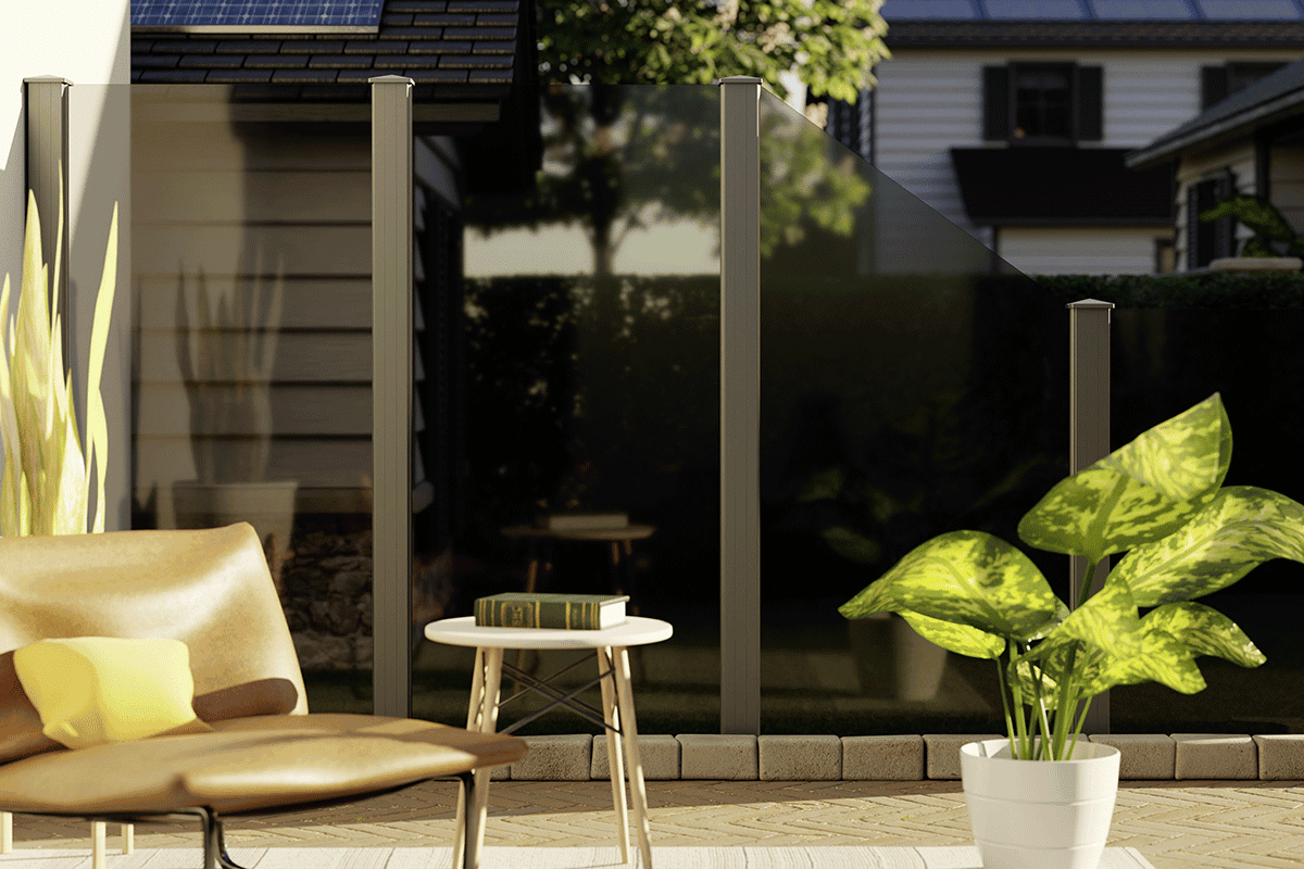 En hyggelig udendørs terrasse har en solbrun loungestol, et lille bord med en bog og en grøn potteplante. Vidual Clear – Glashegn med aluminiumsstolper giver privatliv, mens solpanelklædte huse er synlige i baggrunden.