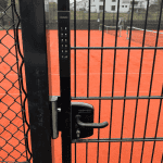 En låst port med Locinox VOLTA - Overflademonteret lås med eksternt batteridrevet tastatur sikrer adgang til en tennisbane af rødt ler; et net og huse ses i baggrunden.