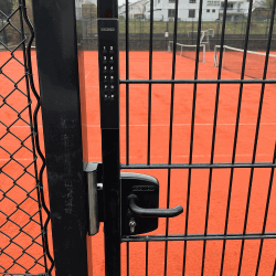 En låst port med Locinox VOLTA - Overflademonteret lås med eksternt batteridrevet tastatur sikrer adgang til en tennisbane af rødt ler; et net og huse ses i baggrunden.