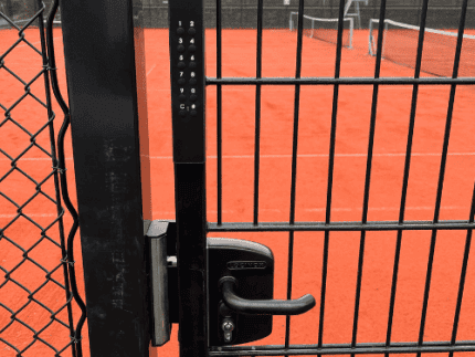 En låst port med Locinox VOLTA - Overflademonteret lås med eksternt batteridrevet tastatur sikrer adgang til en tennisbane af rødt ler; et net og huse ses i baggrunden.