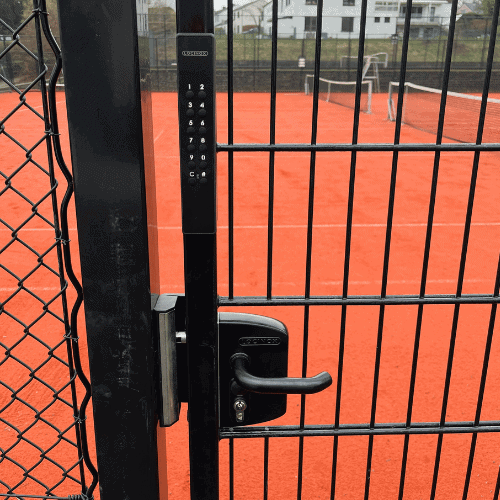 En låst port med Locinox VOLTA - Overflademonteret lås med eksternt batteridrevet tastatur sikrer adgang til en tennisbane af rødt ler; et net og huse ses i baggrunden.