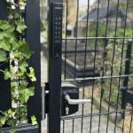 En sort metalport med en Locinox VOLTA overflademonteret lås med eksternt batteridrevet tastatur står delvist åben, omgivet af lodret hegn og grøn efeu. I baggrunden anes en uklar have eller udendørs område.