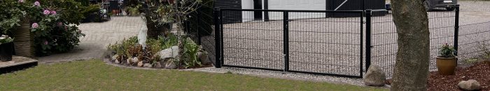 En sort metalport står ved indgangen til en indkørsel med grus, omkranset af planter og sten, der fører til en hvid garageport og bygninger i baggrunden.