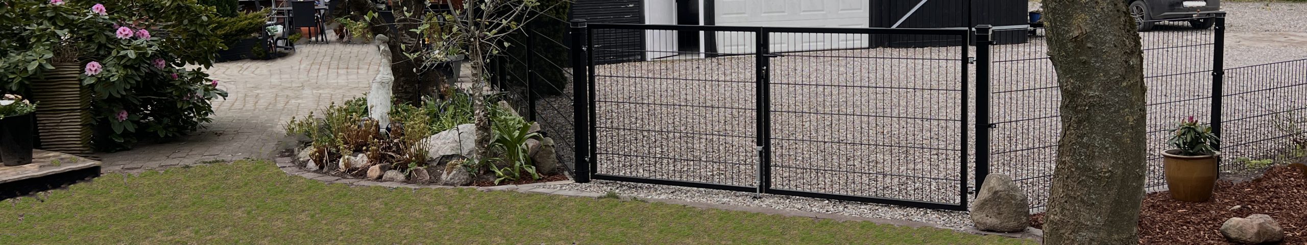 En sort metalport står ved indgangen til en indkørsel med grus, omkranset af planter og sten, der fører til en hvid garageport og bygninger i baggrunden.