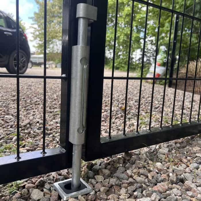 Nærbillede af en Locinox Skyderigel-holder til nedgravning, der fastgør en sort trådport til en jordstik på grus, med en sort bil og grønne områder synlige i baggrunden.