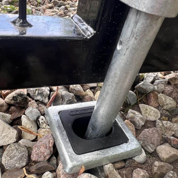 En Locinox Skyderigel-holder til nedgravning, lavet af metal og plastik, er installeret i en firkantet jordfatning omgivet af sten og grus, med et sort metalhegn synligt i baggrunden.