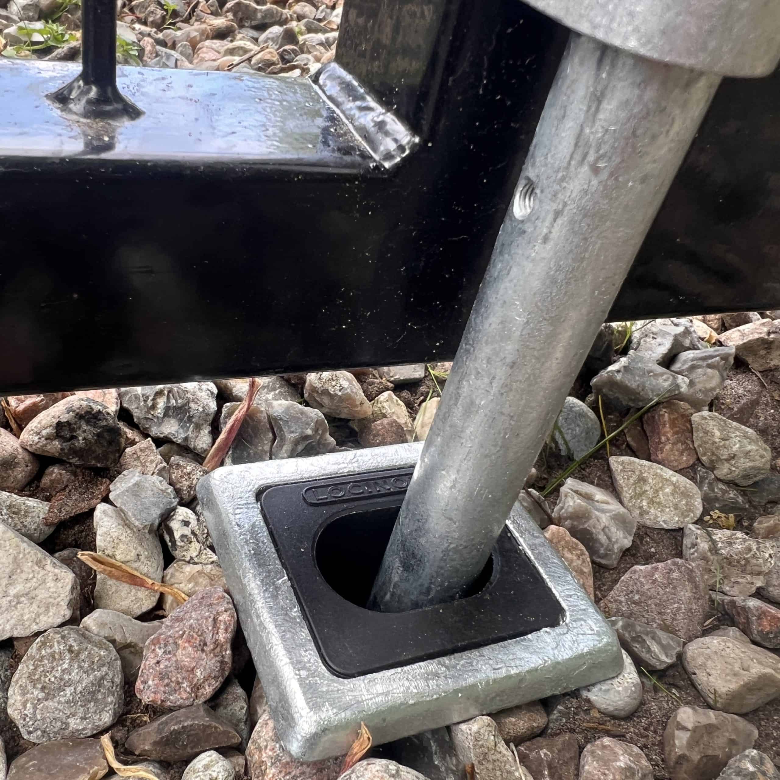 En Locinox Skyderigel-holder til nedgravning, lavet af metal og plastik, er installeret i en firkantet jordfatning omgivet af sten og grus, med et sort metalhegn synligt i baggrunden.