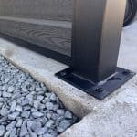 Nærbillede af en sort skraldespandsskærm med kompositfyld – Sort RAL 9005 stolpe, fastgjort til beton med fire bolte ved siden af grå grus – en moderne udendørsløsning med pulverlakerede stolper og u-skinner.