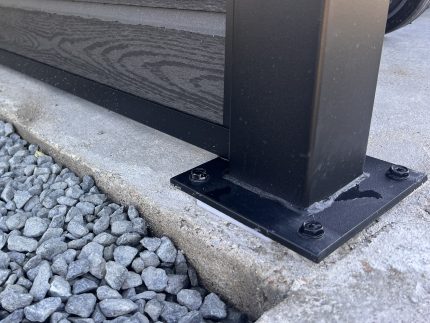 Nærbillede af en sort skraldespandsskærm med kompositfyld – Sort RAL 9005 stolpe, fastgjort til beton med fire bolte ved siden af grå grus – en moderne udendørsløsning med pulverlakerede stolper og u-skinner.