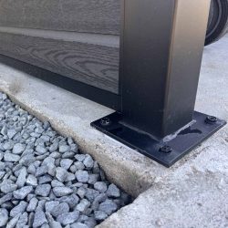 Nærbillede af en sort skraldespandsskærm med kompositfyld – Sort RAL 9005 stolpe, fastgjort til beton med fire bolte ved siden af grå grus – en moderne udendørsløsning med pulverlakerede stolper og u-skinner.