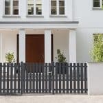 Et moderne hvidt hus med en hoveddør af træ, høje vinduer ovenover og et mørkegråt Classic Line - Aluminium Stakithegn med Lodrette Profiler stakit med låge foran. Grønne potteplanter står på hver side af indgangen.