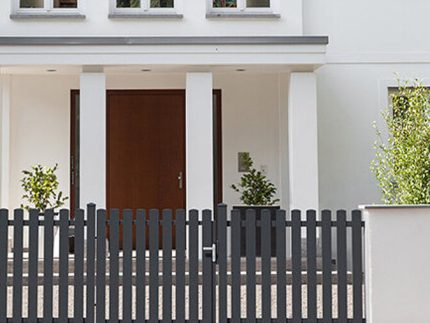 Et moderne hvidt hus med en hoveddør af træ, høje vinduer ovenover og et mørkegråt Classic Line - Aluminium Stakithegn med Lodrette Profiler stakit med låge foran. Grønne potteplanter står på hver side af indgangen.