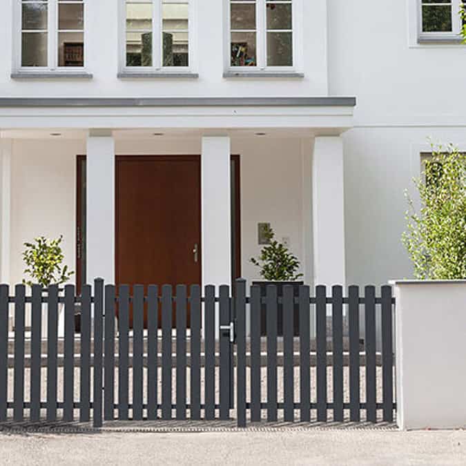 Et moderne hvidt hus med en hoveddør af træ, høje vinduer ovenover og et mørkegråt Classic Line - Aluminium Stakithegn med Lodrette Profiler stakit med låge foran. Grønne potteplanter står på hver side af indgangen.