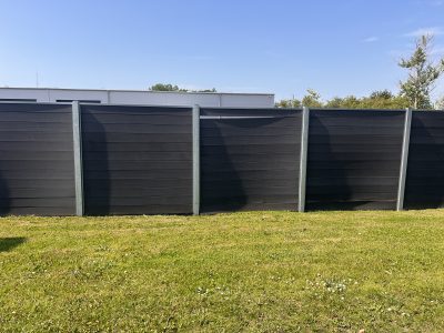 Et sort komposithegn med metalpæle med tynde brædder står på et græsklædt område under en klar blå himmel med træer og en hvid bygning synlig i baggrunden.