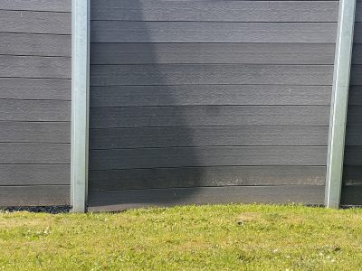 Et nærbillede af et moderne gråt hegn med vandrette paneler, muligvis en komposit hegn monteret forkert, støttet af metalpæle, med grønt græs i forgrunden og en skygge kastet på hegnet.