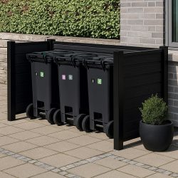 Tre sorte skraldespande står opstillet i en Komposit Skraldespandsskjuler – med sorte pulverlakerede stolper & skinner uden for en murstensbygning. En lille pottebusk står i nærheden med en grøn hæk i baggrunden.