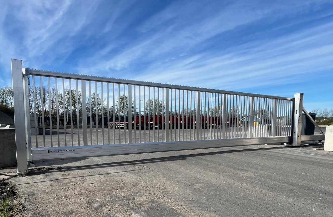 En stor skydeport af metal ses på tværs af en asfalteret indkørsel med træer og industrikøretøjer synlige i baggrunden under en blå himmel med tynde skyer.