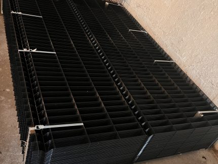 En stak 3D-panelhegn 4/4mm 173 cm SORT (125 m) + stolper 60×40 mm (240 cm) - Outlet-pakke-paneler er pænt organiseret med hvide lynlåse på et betongulv ved siden af en beige struktureret væg.