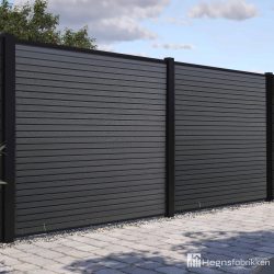 Et moderne Komposit Listeprofil 25mm - WPC Træstruktur hegn med mørkegrå vandrette lameller og sorte stolper står på en asfalteret flade med grønne områder bagved. Hegnsfabrikkens logo er synligt i nederste højre hjørne.