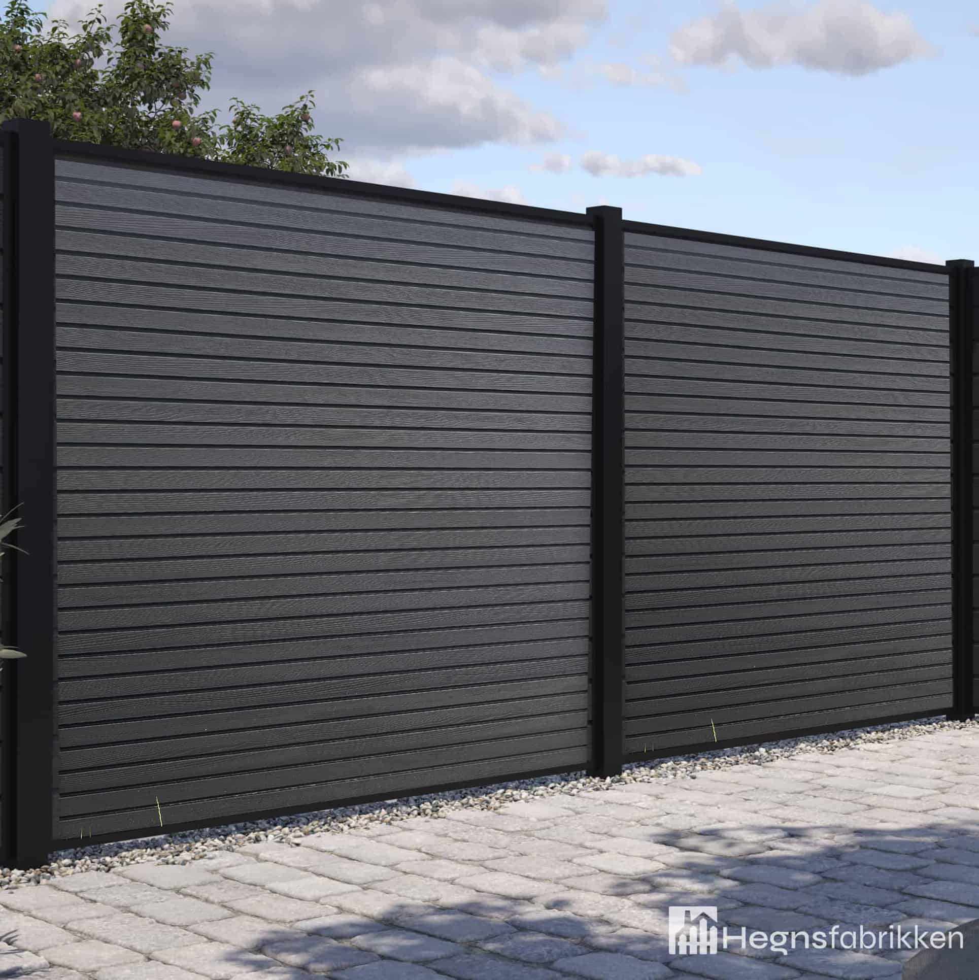 Et moderne Komposit Listeprofil 25mm - WPC Træstruktur hegn med mørkegrå vandrette lameller og sorte stolper står på en asfalteret flade med grønne områder bagved. Hegnsfabrikkens logo er synligt i nederste højre hjørne.