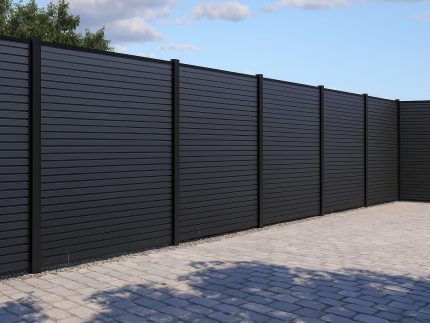 Et moderne sort metalhegn med Komposit Listeprofil 25 mm - WPC Træstruktur-lameller omslutter en brolagt gårdsplads med træer og en delvis overskyet himmel i baggrunden.