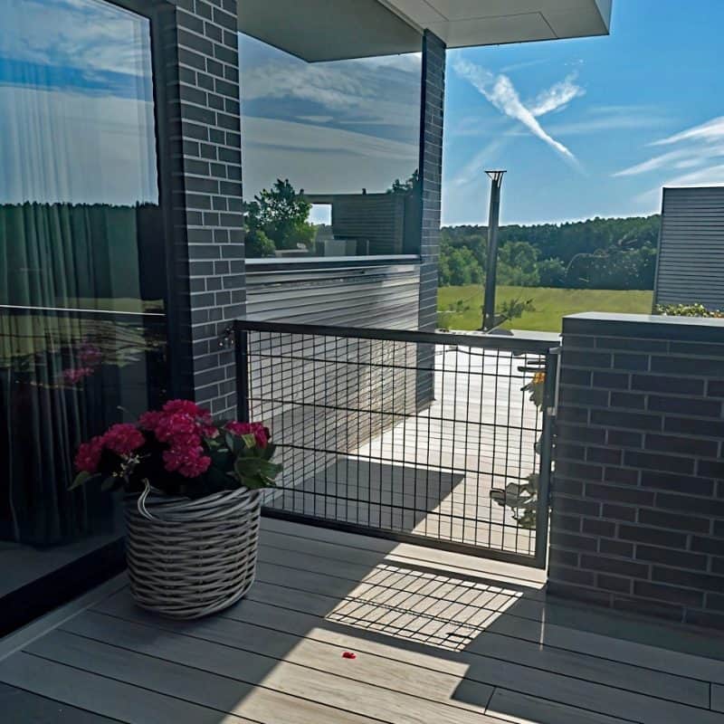En solrig altan med en metalport i pulverlakeret stål, omgivet af mørke murstensvægge. Ved glasdøren står en potteplante med lyserøde blomster, og i baggrunden ses grønne træer og en blå himmel med skyer.