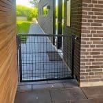 En sort metalport i pulverlakeret stål med gittermønster står mellem to moderne murstens- og trævægge og åbner ud til en brolagt gårdhave med grønt græs og træer i baggrunden.