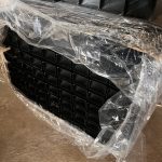 En sort plastbakke med flere firkantede celler, delvist pakket ind i klar plast, står på beton ved siden af en 3D-panelhegn 4/4mm 173 cm SORT (125 m) + stolper 60×40 mm (240 cm) - Outlet-pakke.