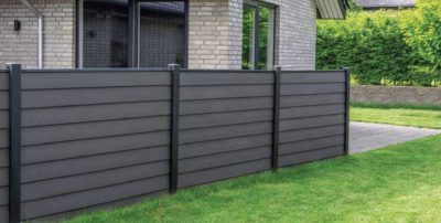 Et moderne hus med lyse murstensvægge og store vinduer er afgrænset af et vandret mørkegråt HF dobbeltklink kompositprofiler med træstruktur - 25 mm hegn og ligger på en pænt trimmet grøn græsplæne med en hæk i baggrunden.