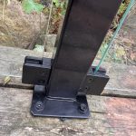 Et nærbillede af Glasholder firkantet model 23P - sort RAL 9005 monteret på forvitret træ, der understøtter klare glaspaneler, med grønne planter og klipper i baggrunden.