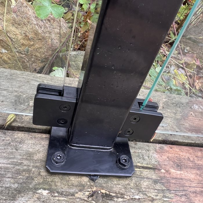 Et nærbillede af Glasholder firkantet model 23P - sort RAL 9005 monteret på forvitret træ, der understøtter klare glaspaneler, med grønne planter og klipper i baggrunden.