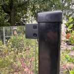 Et nærbillede af Glasholder firkantet model 23P - sort RAL 9005, der fastgør et glashegnspanel til en sort stolpe med grønne områder, træer og en stenstatue i baggrunden.