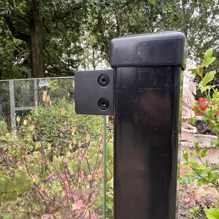 Et nærbillede af Glasholder firkantet model 23P - sort RAL 9005, der fastgør et glashegnspanel til en sort stolpe med grønne områder, træer og en stenstatue i baggrunden.