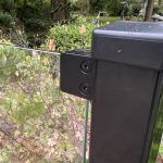 Nærbillede af en Glasholder firkantet model 23P - sort RAL 9005, der fastgør et glashegnspanel til en sort stolpe med to skruer, omgivet af frodige blade og planter i haven.