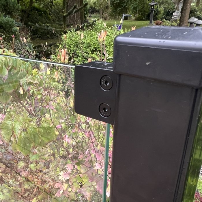 Nærbillede af en Glasholder firkantet model 23P - sort RAL 9005, der fastgør et glashegnspanel til en sort stolpe med to skruer, omgivet af frodige blade og planter i haven.