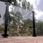 Glasholder firkantet model 23P - sort RAL 9005, et klart glashegn med sorte metalstolper, er installeret på et trædæk med træer og buske i baggrunden under en overskyet himmel.