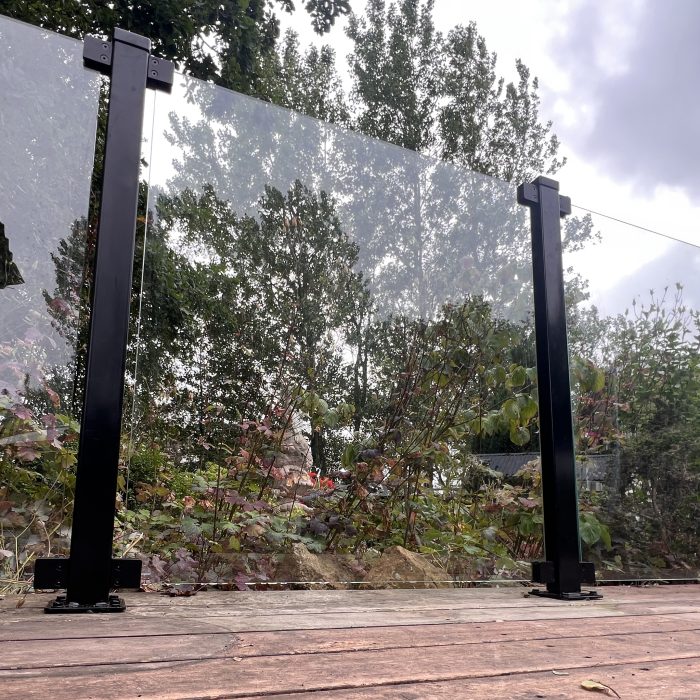 Glasholder firkantet model 23P - sort RAL 9005, et klart glashegn med sorte metalstolper, er installeret på et trædæk med træer og buske i baggrunden under en overskyet himmel.